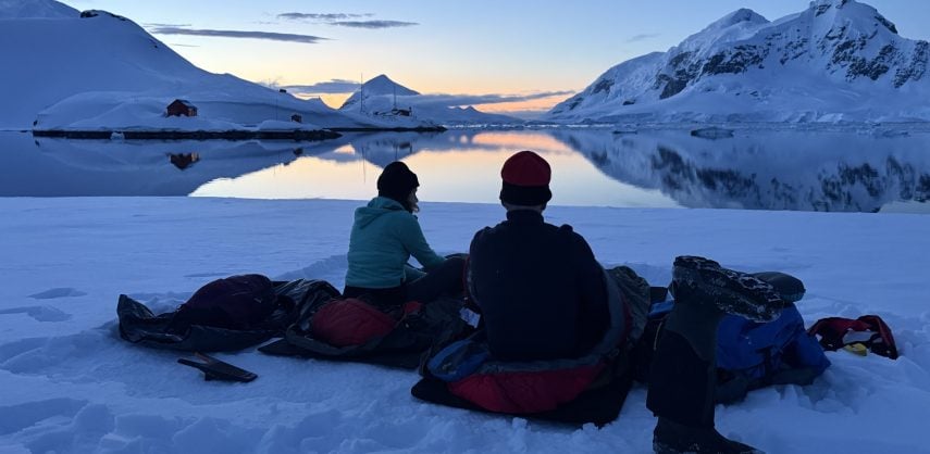 Two people in sleeping bags in the snow, looking out over the coast of the Antarctic Peninsula at sunset