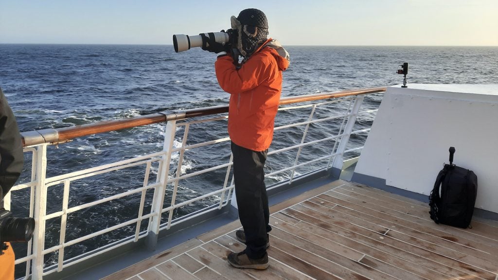 A man in an orange parka and warm hat holds a large camera. He is standing on the deck of an expedition cruise ship in the Southern Ocean, and is looking down his camera for seabirds