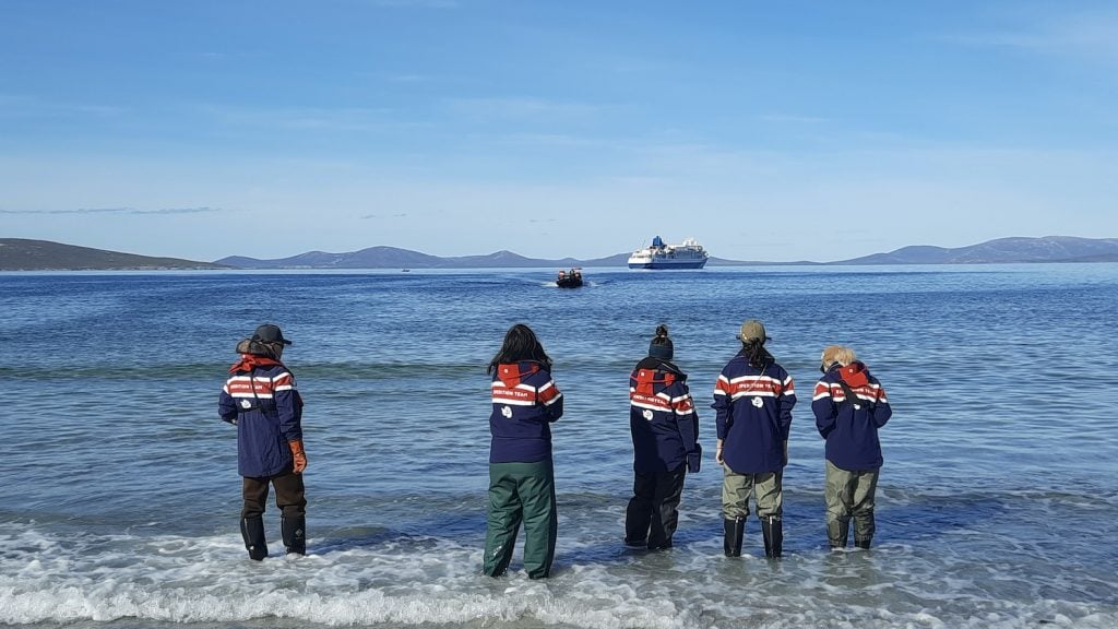 Five expedition guides stand on a beach, waiting for a zodiac full of cruise passengers to land in the Falkland Islands. The cruise ship Seaventure can be seen in the distance.