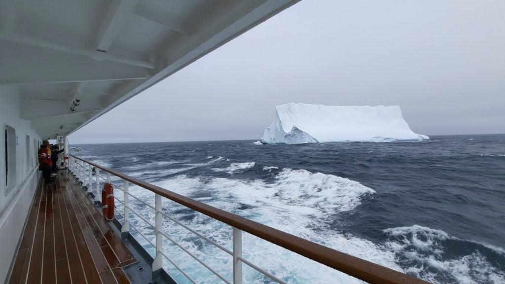 On a cloudy day, an enormous iceberg is spotted from the side deck of an expedition cruise ship.