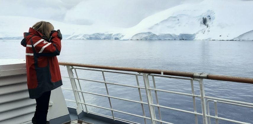 A passengers in a red coat stands on the deck of an expedition cruise ship taking a photo of any icy Antarctic view