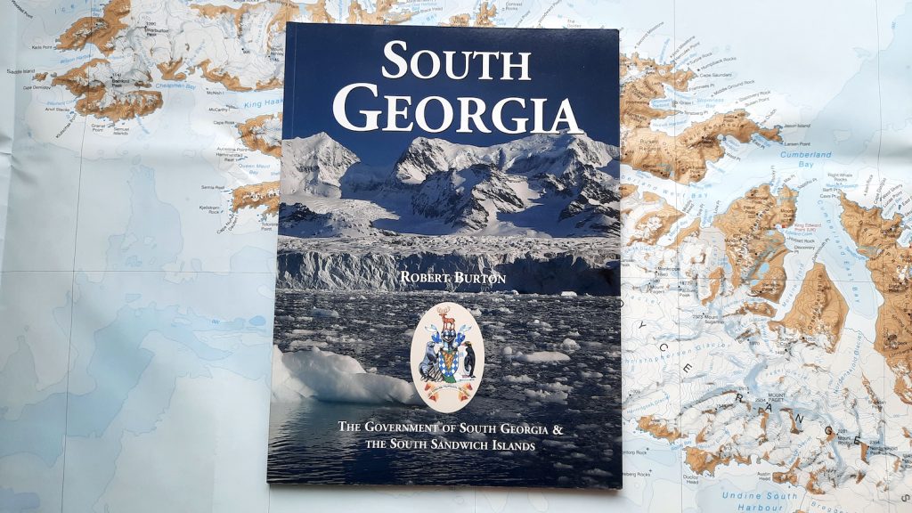 A copy of the official visitor's handbook to South Georgia by the Government of South Georgia & the South Sandwich Islands, and written by Robert Burton. The book sits on background of a map of South Georgia