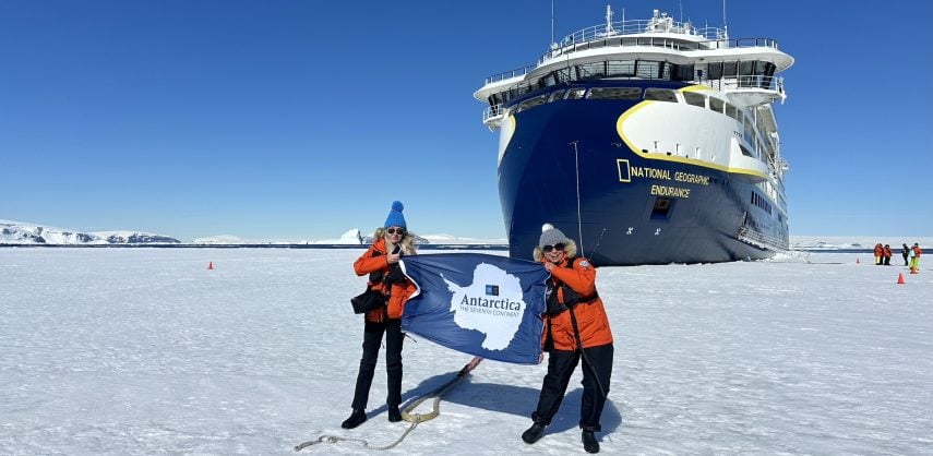 Two tourists on the Antarctic ice holding a flag with the ship National Geographic Endurance in the background